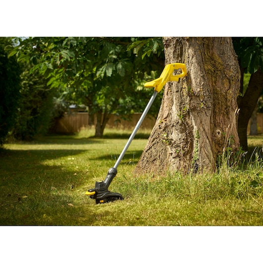STANLEY SFMCST925M1 grass trimmer with yellow handle resting on tree in garden.