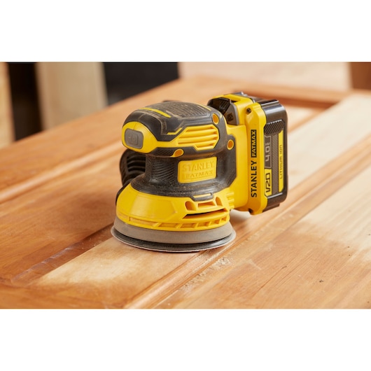 STANLEY SFMCW220M1S cordless random orbital sander in yellow and black, sanding wooden surface, close-up.