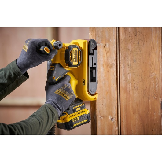 STANLEY SFMCW223B-XJ cordless sander in use on wood, close-up of yellow tool held by gloved hands.