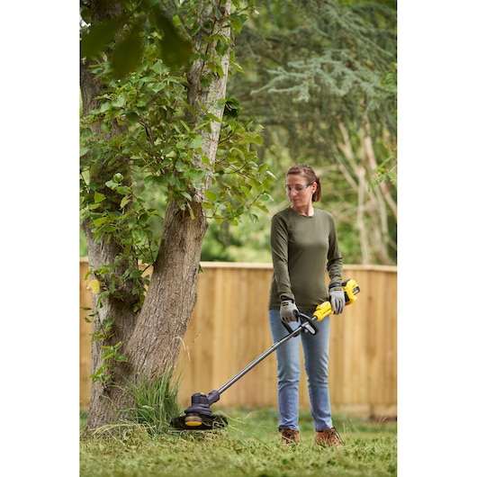 Photo of person using STANLEY SFMSTB930M grass trimmer outdoors near a tree, wearing gloves and casual clothing.