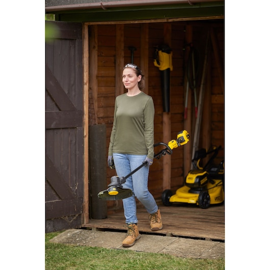 STANLEY SFMSTB930M grass trimmer held by a person in front of a shed, lawnmowers visible inside.