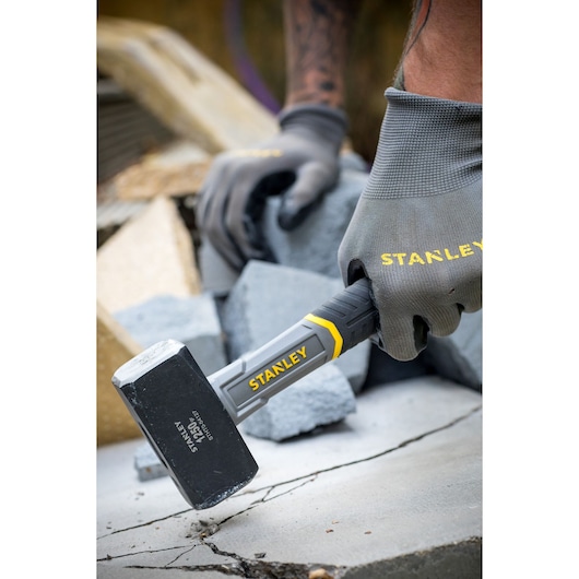 Photo of STANLEY STHT0-54127 club hammer in use, black head with yellow accents, held by gloved hand on concrete.