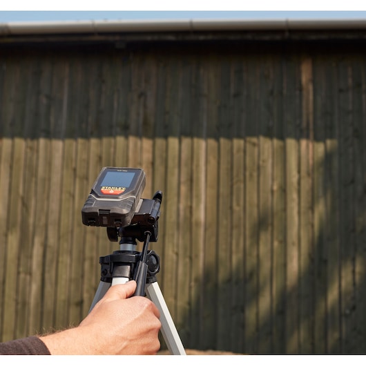 Photo of STANLEY STHT1-77142 laser distance measurer on tripod, hand adjusting device outdoors.