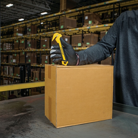 Photo of a gloved hand using a STANLEY STHT10356 safety box cutter to open a cardboard box in a warehouse.