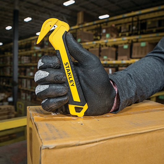 Photo of STANLEY STHT10356 yellow safety box cutter in gloved hand opening cardboard box in warehouse.