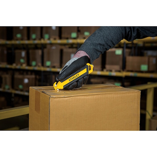 Photo of STANLEY STHT10360-1 safety utility knife cutting cardboard box in warehouse setting.