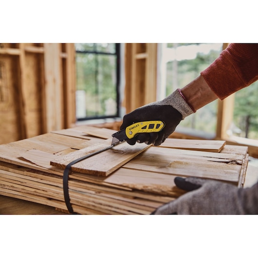 STANLEY STHT10424 utility knife with yellow and black handle in use cutting strap on stack of wooden boards.