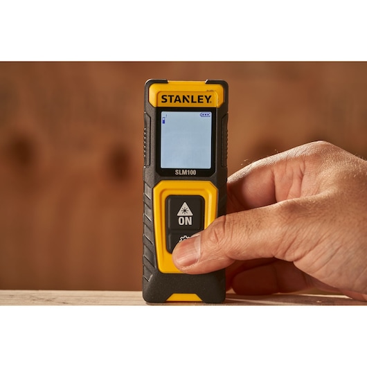 Stanley STHT77100-0 laser distance measurer handheld, front view, with thumb pressing ON button.