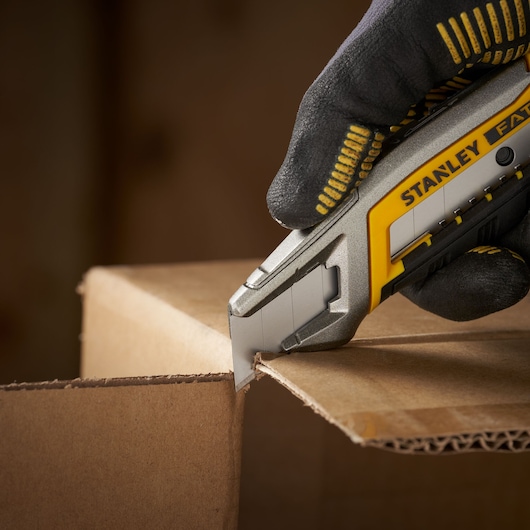 Photo of STANLEY STHT9-11301 utility knife cutting cardboard, held by gloved hand, close-up view.
