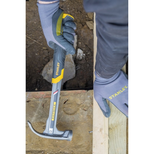 STANLEY XTHT1-51148 claw hammer in use, silver with yellow handle, shown nailing wood, person wearing gloves.